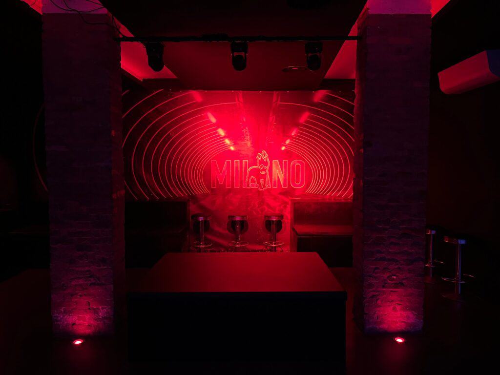 Illuminazione rossa e neon con testo Milano e logo al centro, atmosfera vivace e accogliente.